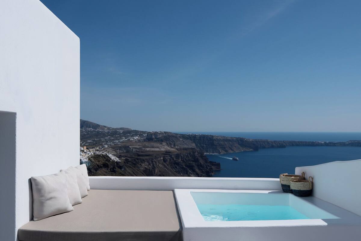 Un balcon doté d’un espace salon avec des coussins blancs et d’une petite piscine, offrant une vue sur un paysage côtier avec des falaises et la mer.