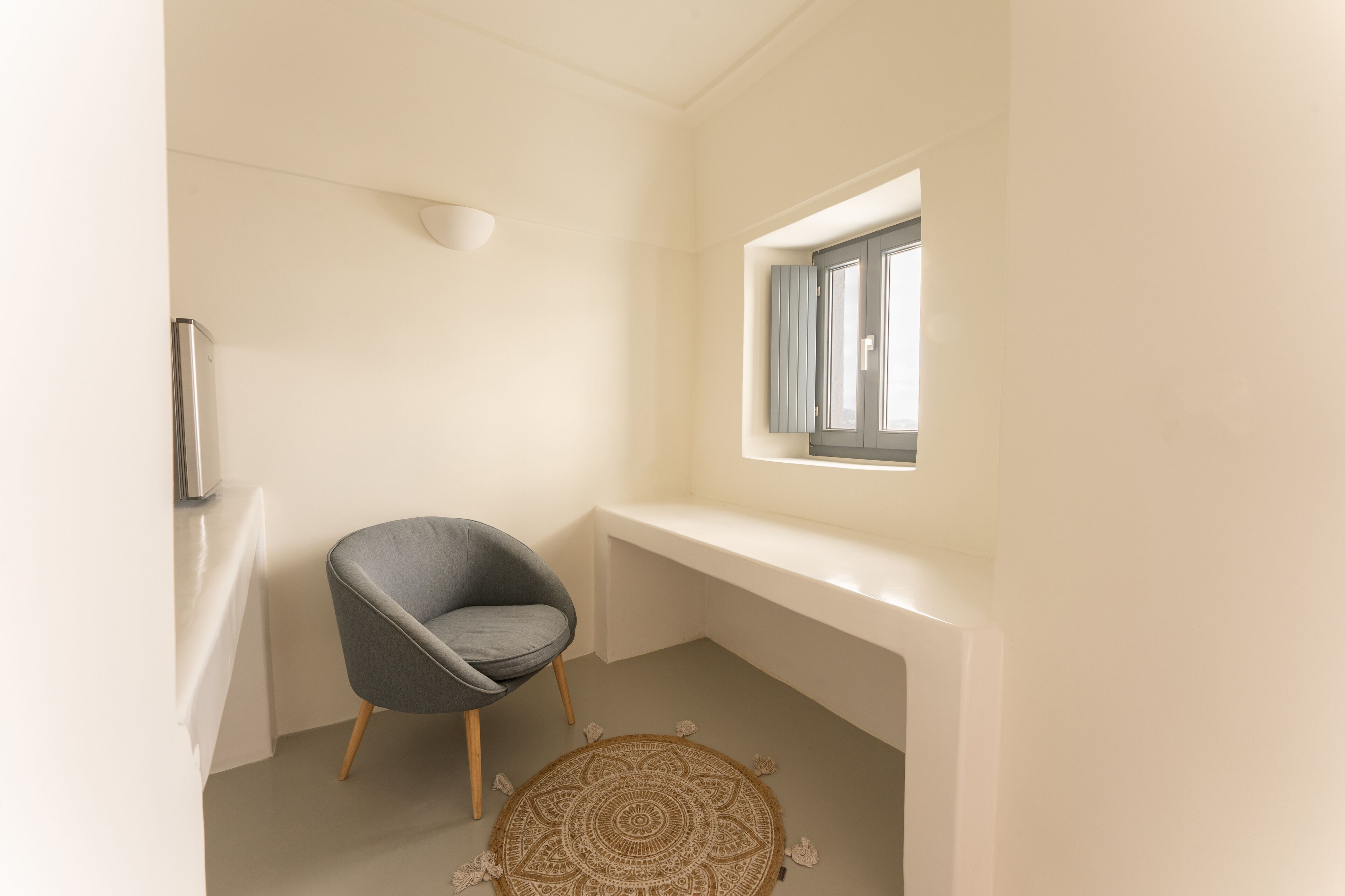 A small, modern room with a single gray chair, a round patterned rug, and a window with blinds.