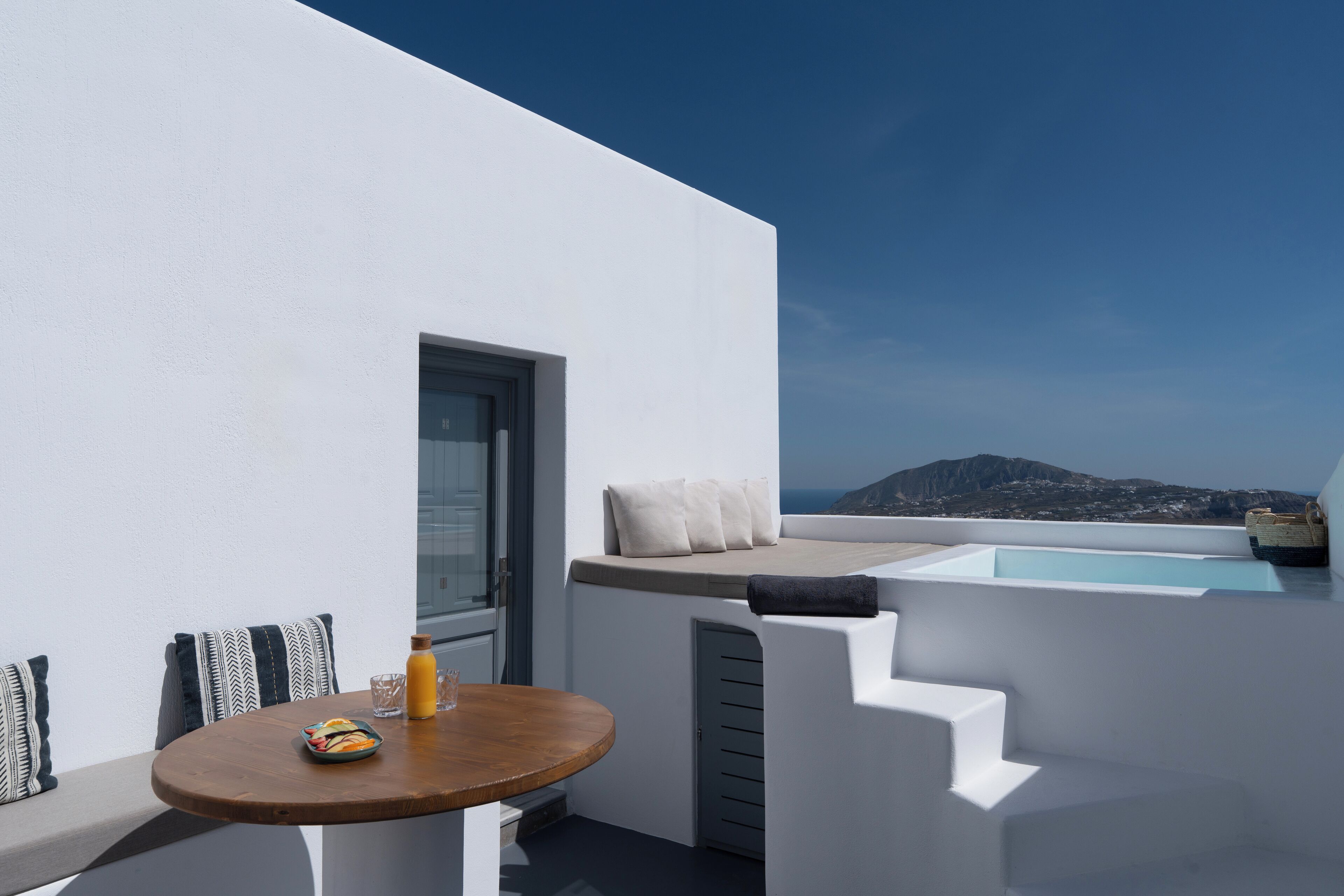 A modern outdoor seating area with a table, cushions, and a view of the sea and mountains.