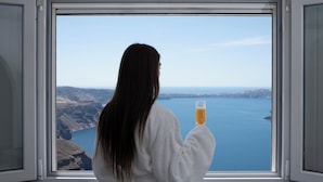 Una persona con bata blanca sosteniendo un vaso de jugo de naranja, mirando por una ventana con vista a una gran masa de agua y tierra distante.