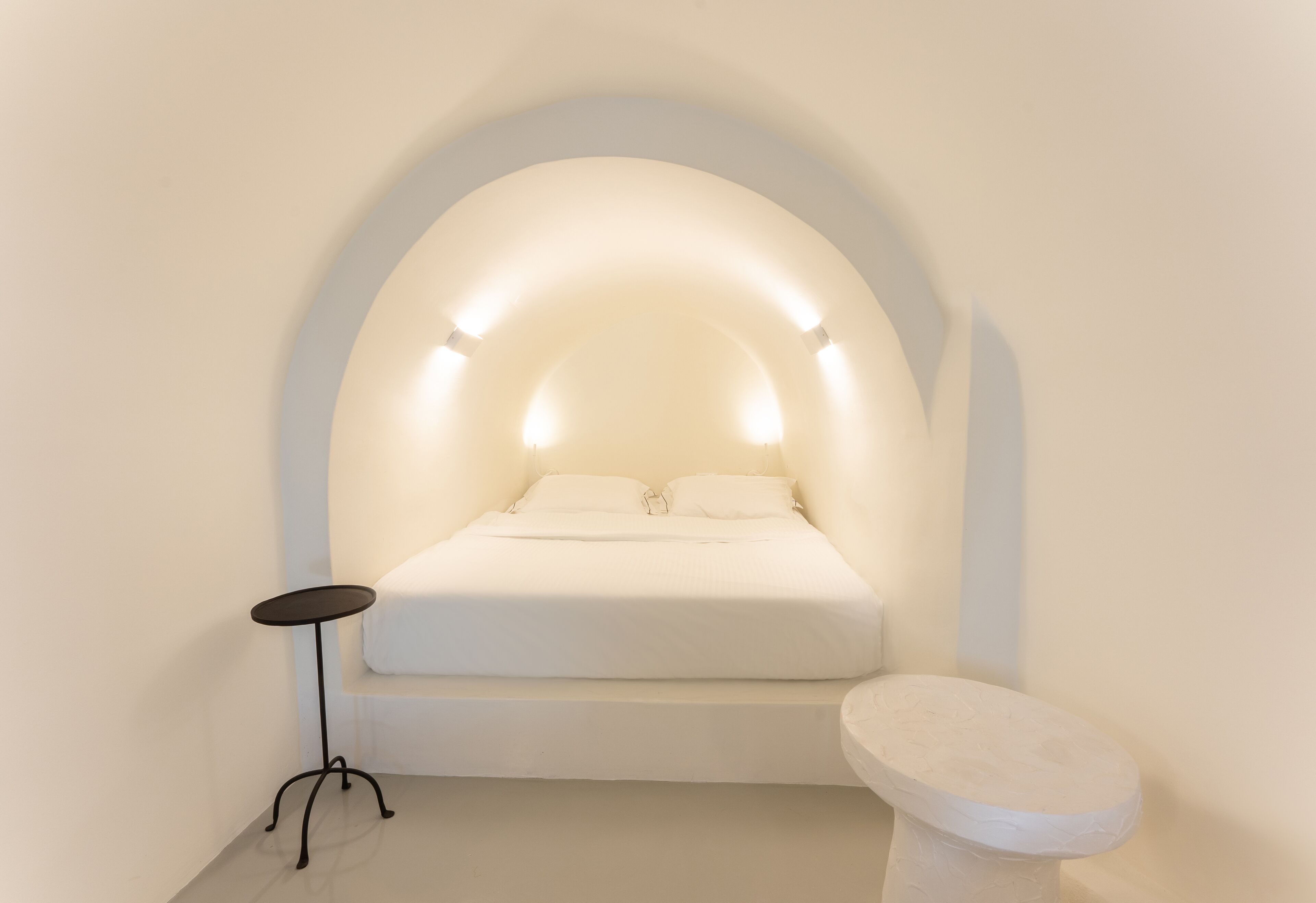 A minimalist bedroom with a bed under an arched ceiling, a small round table, and a toilet.