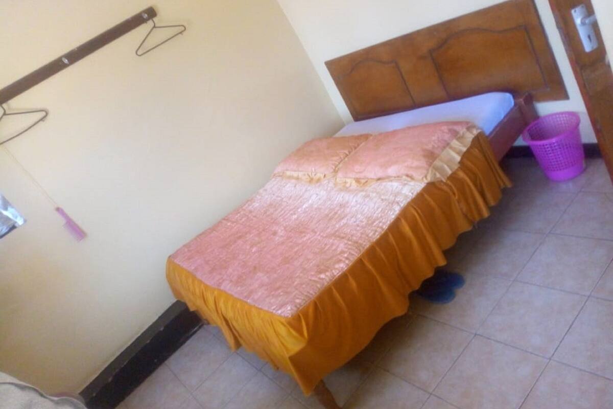 A small, single-bed room with a wooden headboard, a bed with a pink and orange bedspread, a purple laundry basket, and a wall-mounted hanger.