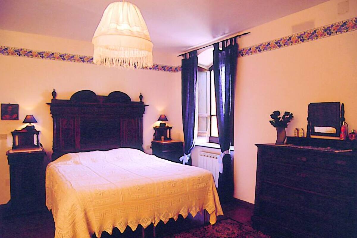 Une chambre à coucher avec un grand lit, deux tables de chevet, une commode, un lustre et une fenêtre avec des rideaux.