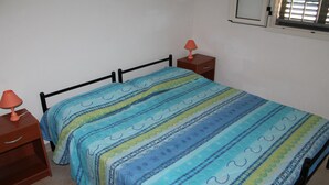 Een eenpersoonsbed met een gestreept dekbedovertrek, een nachtkastje met een lamp en een raam met jaloezieën.