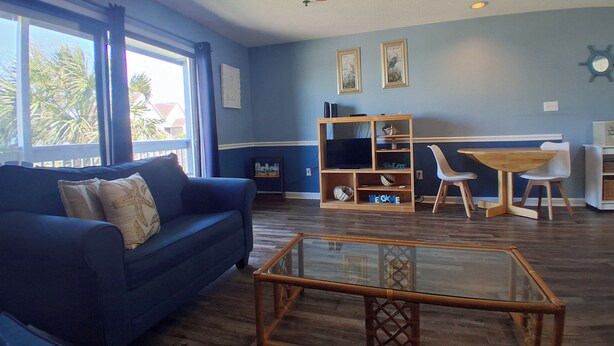 Een woonkamer met een blauwe bank, een glazen salontafel, een houten tv-meubel en een eethoek met een witte tafel en stoelen.