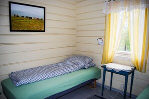 Quarto pequeno com uma cama de solteiro, colcha verde, quadro na parede, mesa de cabeceira com abajur e janela com cortinas amarelas.
