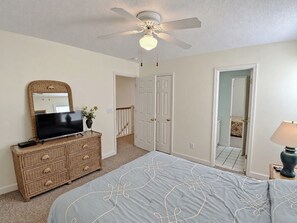 A bedroom with a bed, dresser, television, ceiling fan, and a door leading to another room.