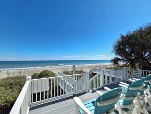 A deck with white railings overlooking a beach with waves and a clear sky.