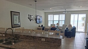 A kitchen with granite countertops, a dining area with white chairs, a TV mounted on the wall, and a view of the ocean through large windows.