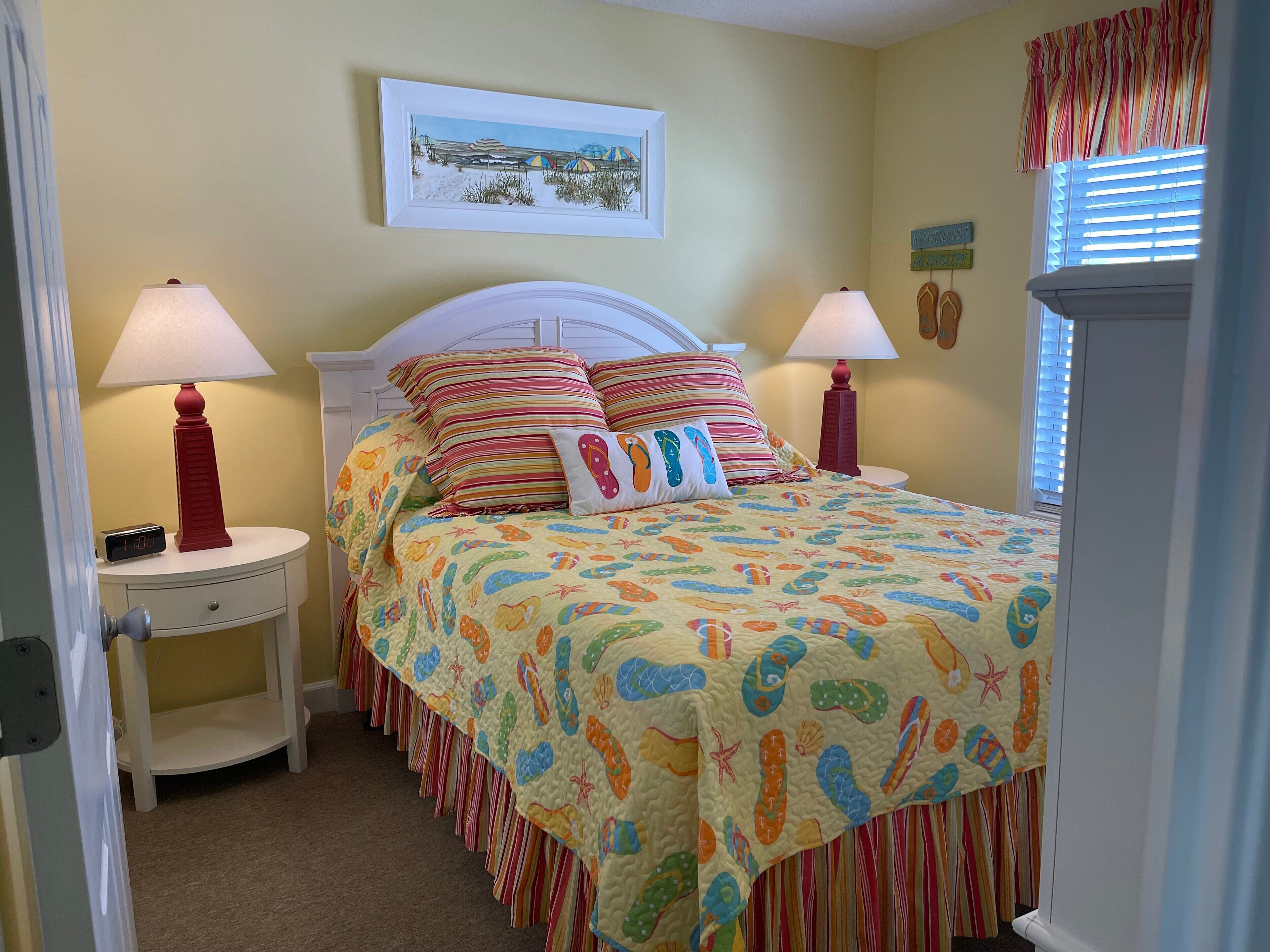 A bedroom with a bed, two bedside tables, a nightstand, a wall clock, and a window with blinds.