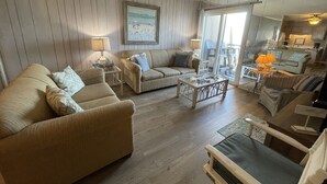 A spacious living room with a large sofa, a smaller sofa, and a coffee table. There is a dining area with a table and chairs visible through a glass door. - Inlet Pointe 37 - Second Row (Channelfront) in Garden City Beach (Garden City)