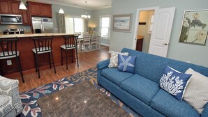 A modern kitchen with stainless steel appliances, a dining area with a chandelier, a blue sofa with decorative pillows, and a patterned rug. - Just Beachin' - Second Row complex in Garden City Beach (Garden City)
