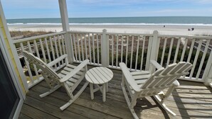 Terraza de madera con sillas mecedoras blancas y una mesita, con vista a la playa.