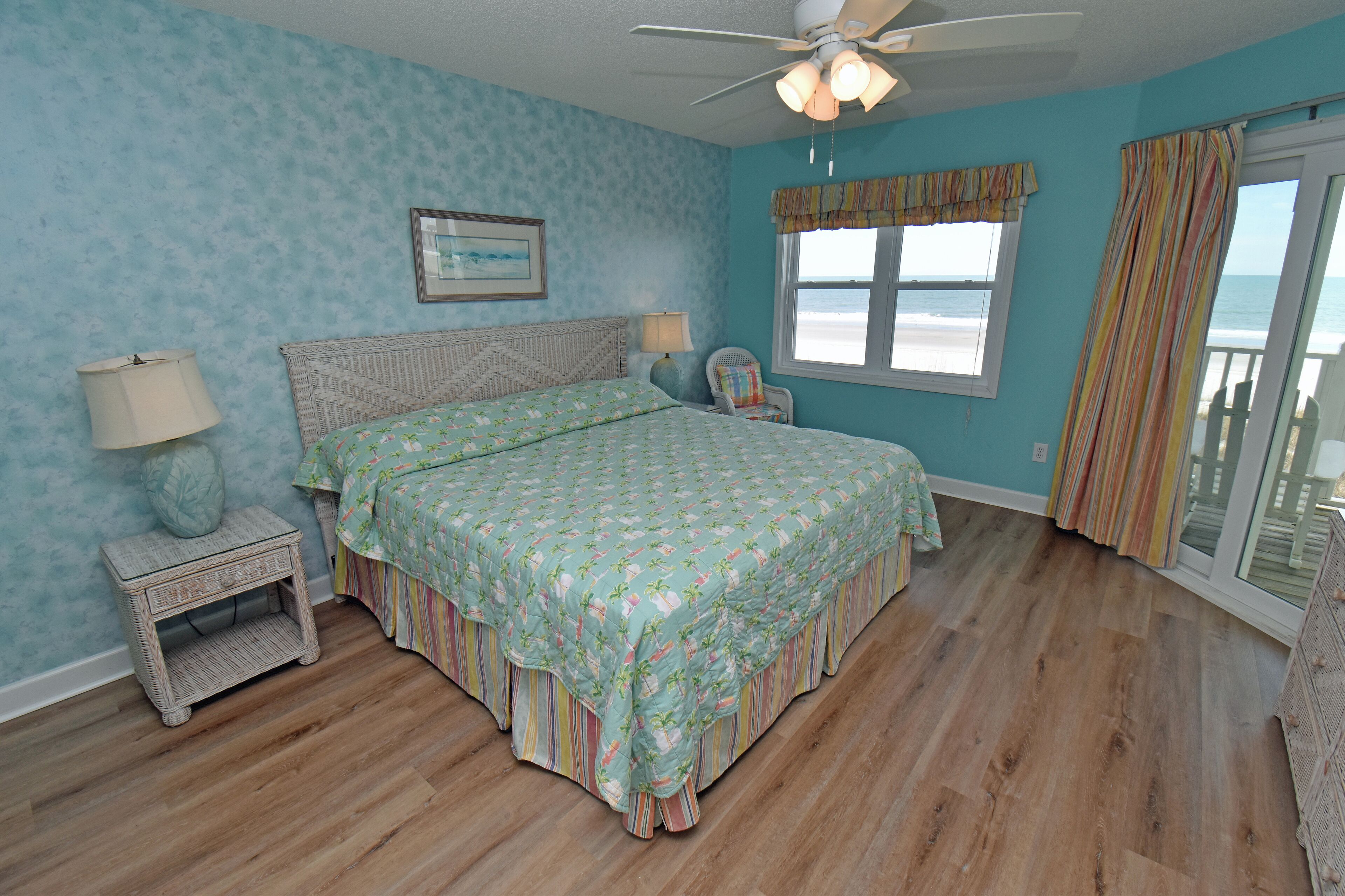 A bedroom with a bed, a nightstand, a chair, a window with a view of the beach, and a ceiling fan.