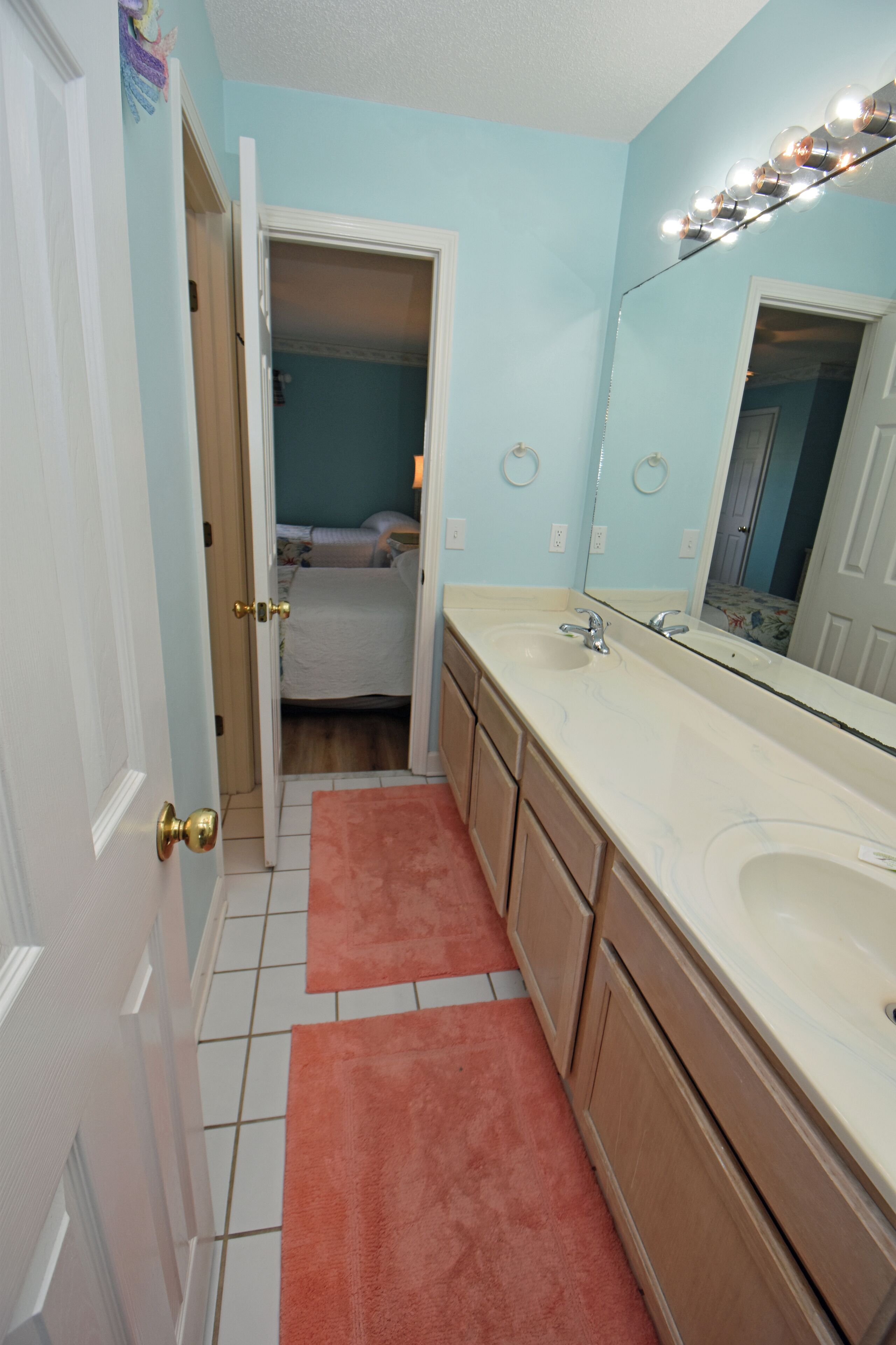 A bathroom with a double sink, a mirror, and a door leading to a bedroom.