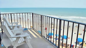Un balcon avec des chaises blanches donnant sur une plage parsemée de parasols et où se trouvent des gens.