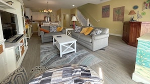 Un salon spacieux avec un canapé gris, une table basse blanche et un tapis à motifs. Un escalier mène à l’étage supérieur.