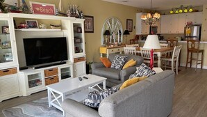 Un salon avec un canapé gris, une table basse blanche et un téléviseur posé sur un meuble. On y trouve également un coin repas avec une table et des chaises, et une cuisine à l’arrière-plan.
