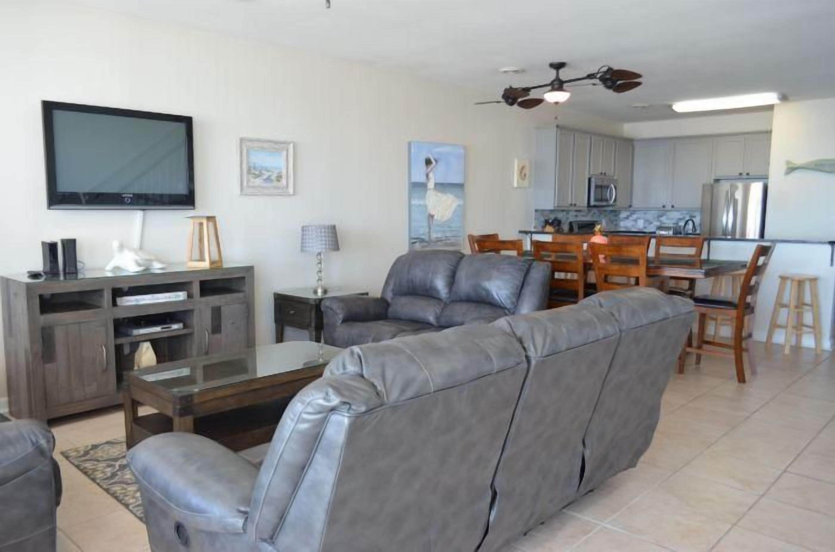 A modern living room with a gray leather sofa, a flat-screen TV, a wooden coffee table, and a dining area with wooden chairs and a kitchen in the background.