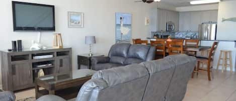 A modern living room with a gray leather sofa, a flat-screen TV, a wooden coffee table, and a dining area with wooden chairs and a kitchen in the background.