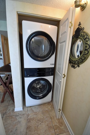 Una sala de lavandería con lavadora y secadora de carga frontal, una silla de madera y un espejo decorativo en la pared.