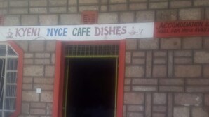 Entrada de un edificio con un letrero que dice 'KYENI NYCE CAFE DISHES' y otro letrero que indica 'ACCOMODATION & HALL, FOR HIRE AVAILABLE'.