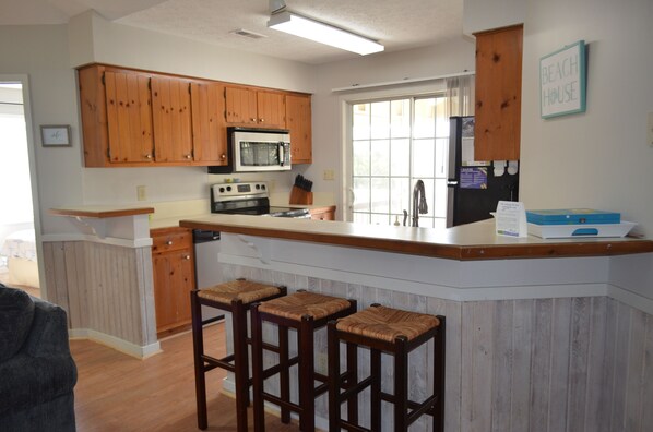 Cocina con armarios de madera, microondas, estufa y refrigerador. Hay tres taburetes frente a la encimera.