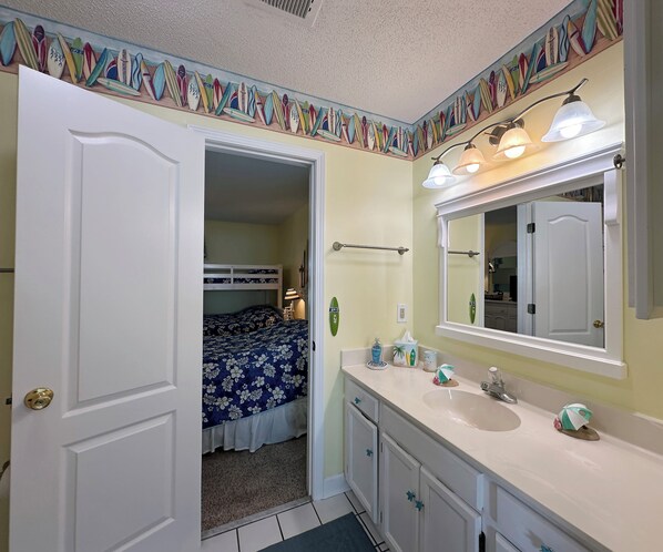 A bathroom with a white vanity, a large mirror, and a door leading to a bedroom with a bunk bed. - Portofino I 1416 - Oceanfront in Garden City Beach (Garden City)
