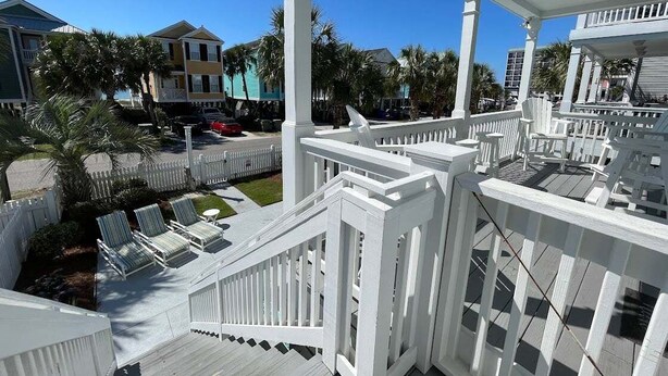 Una terraza de madera blanca con baranda blanca, un pequeño espacio exterior con sillas y vistas a una zona residencial con casas y palmeras.