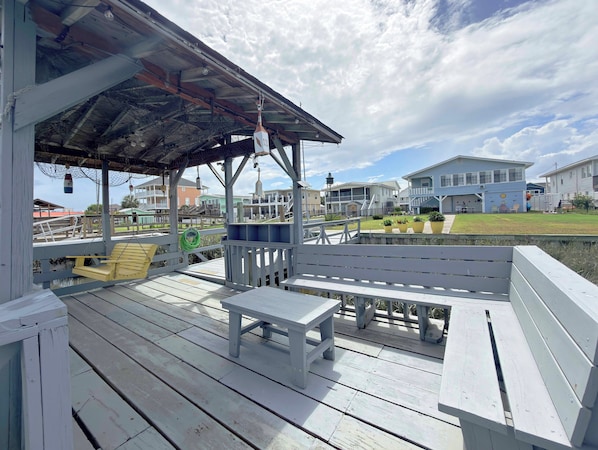 A wooden deck with a bench and table, a covered area with hanging lights, and a view of houses in the background. - Suit Up - Walk to Beach (Channelfront) in Garden City Beach (Garden City)