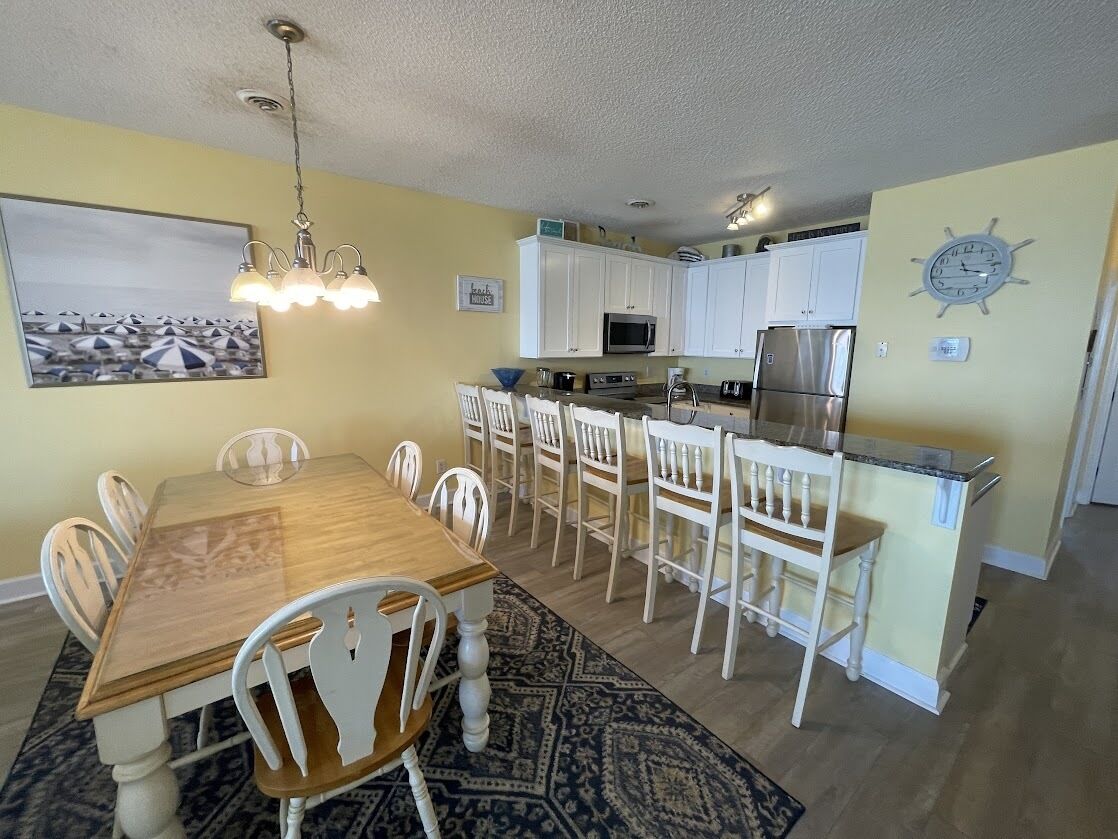 A dining area with a wooden table and white chairs, a kitchen with white cabinets and stainless steel appliances, and a patterned rug on the floor.
