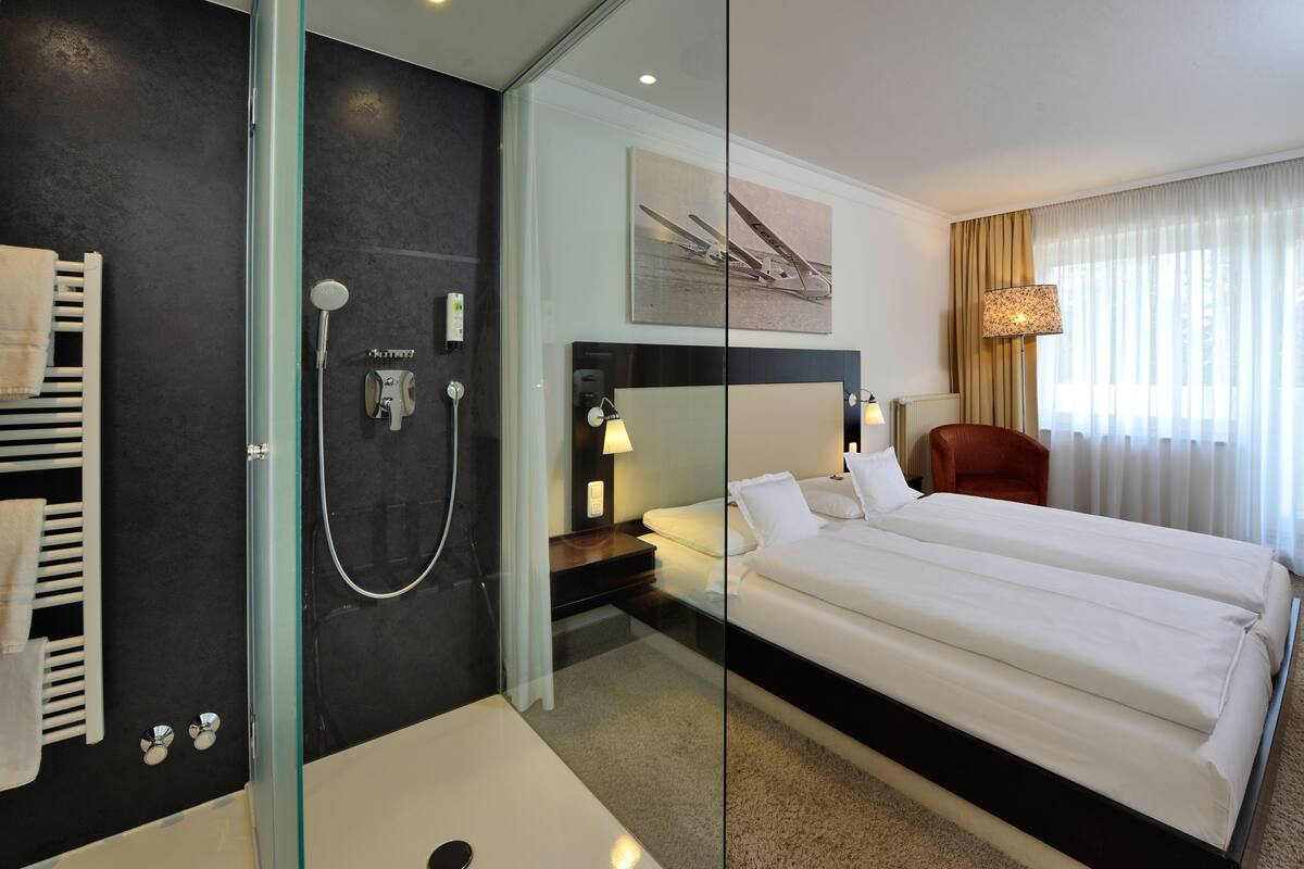 Ein modernes Hotelzimmer mit einer gläsernen Dusche, einem Doppelbett, einem Nachttisch mit Lampe und einem großen Fenster mit Vorhängen.