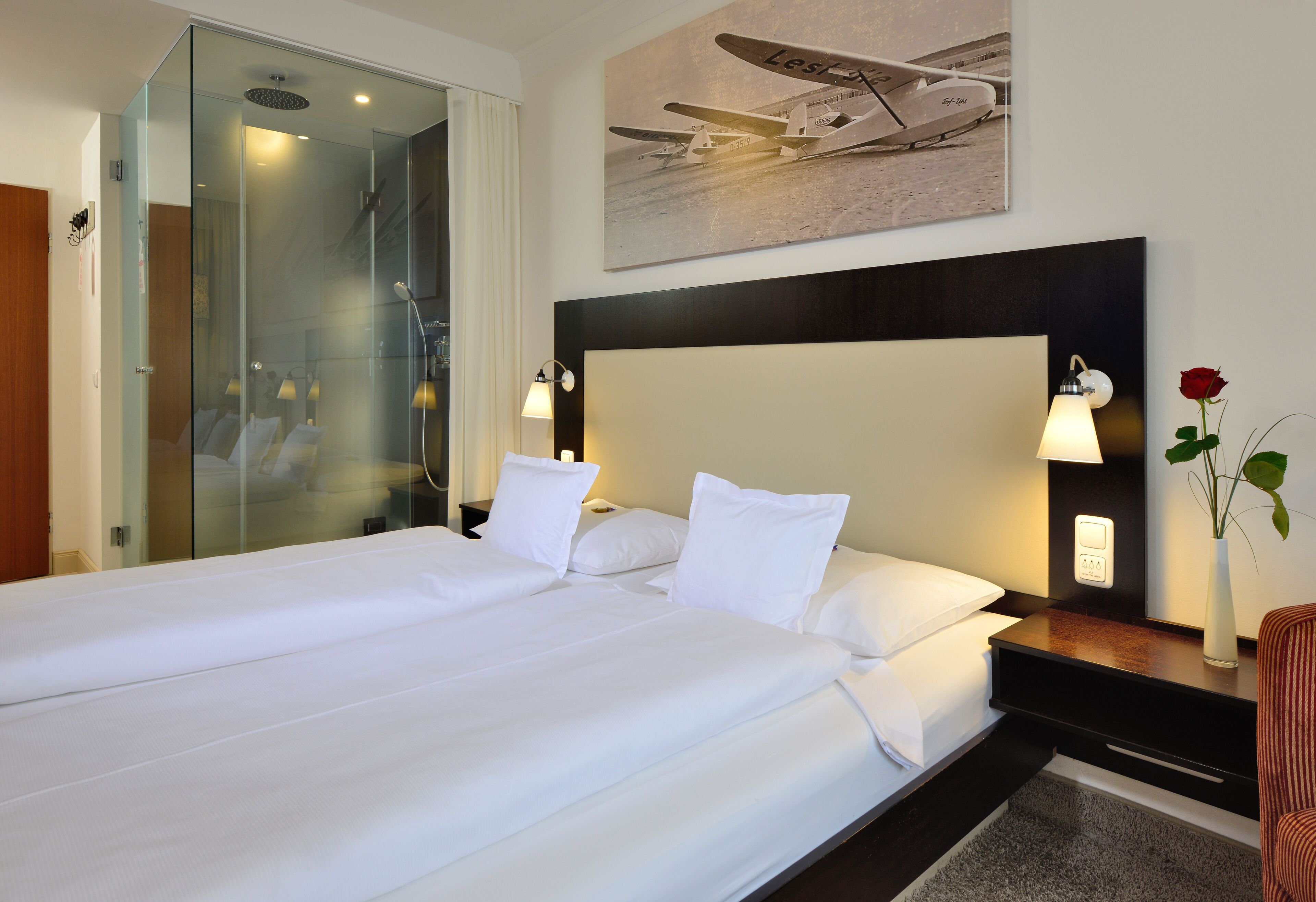 Habitación de hotel con una cama grande, cabina de ducha de vidrio, un cuadro de un avión y una jarra con una sola rosa roja.