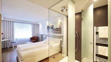A modern hotel room with a glass-enclosed shower, a bed, a desk, and a chair.
