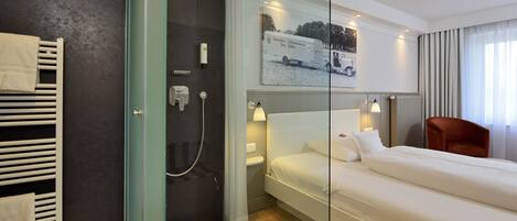 A modern hotel room with a glass shower enclosure, a bed with white linens, a red armchair, and a wall-mounted artwork of a vintage car.