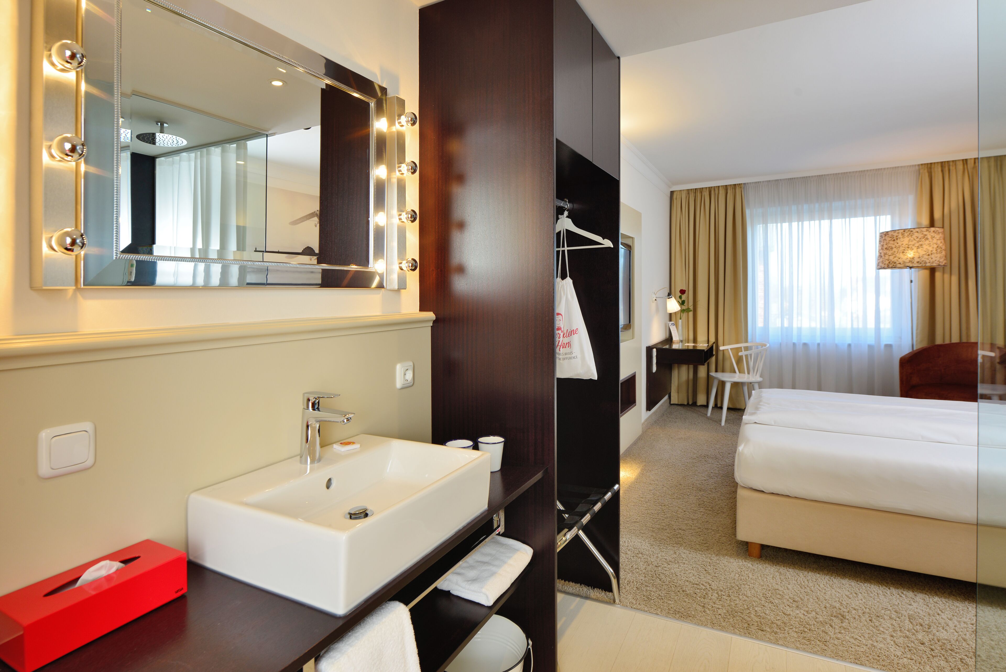 Habitación de hotel con un espejo grande, tocador con lavabo, cama, silla y lámpara.