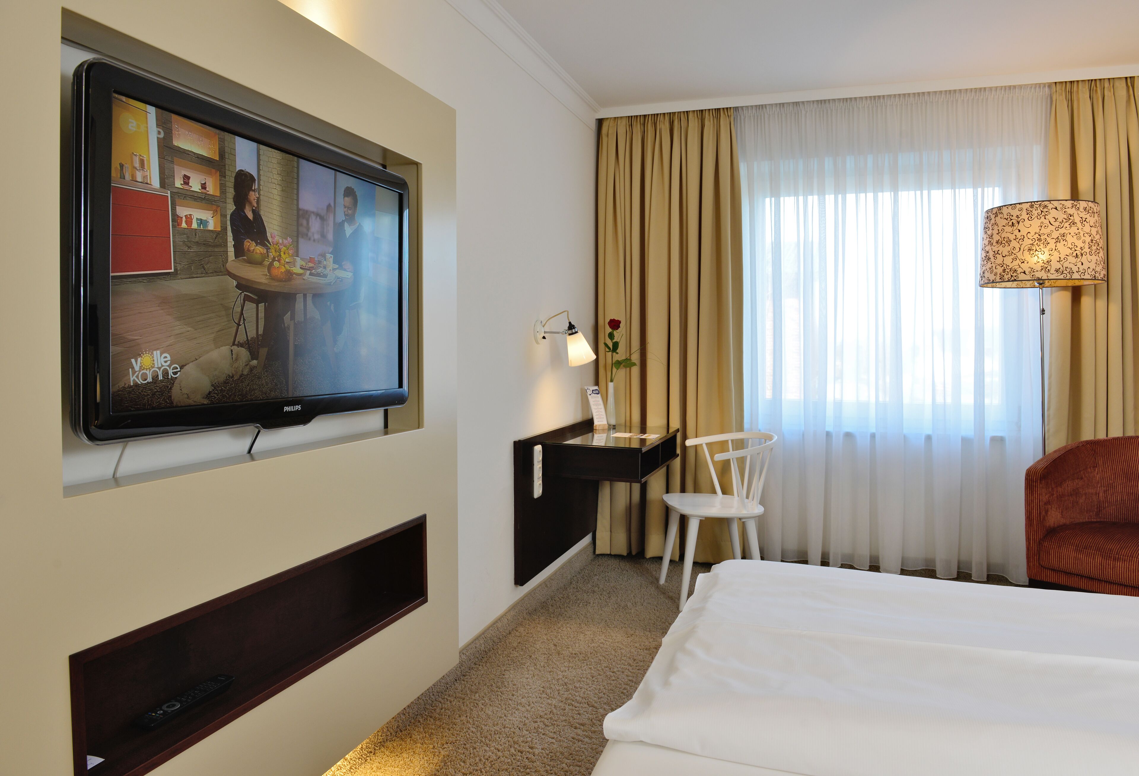 A hotel room with a television mounted on the wall, a desk with a lamp, a chair, a bed, and a window with curtains.