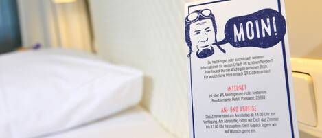Camera d'albergo con un letto, un comodino e un cartello con informazioni su Wi-Fi e disponibilità delle camere.