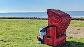 A red wicker chair with a black strap, placed on a grassy field with a view of the sea and a distant ship in the background.
