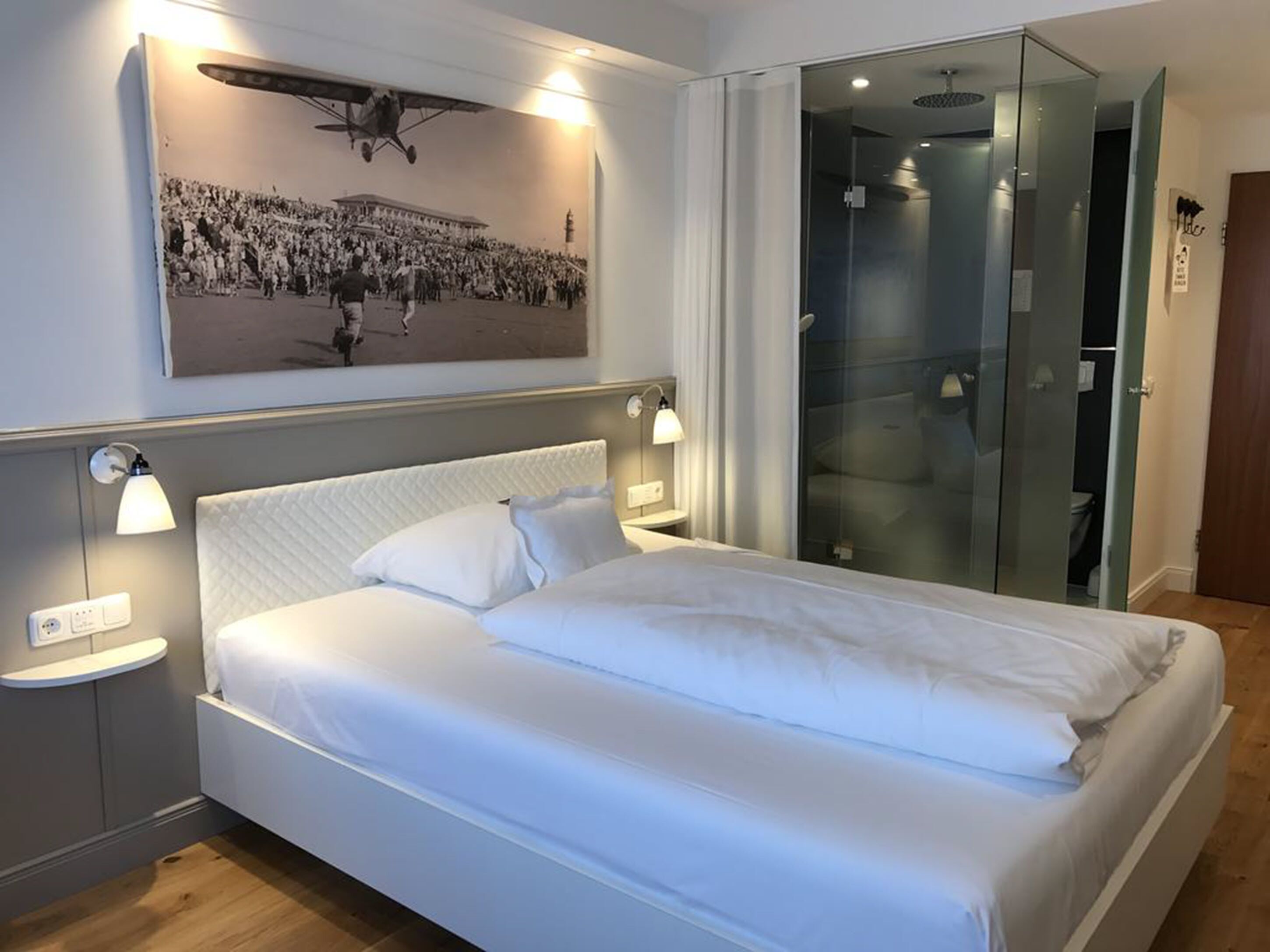 Una habitación de hotel moderna con una cama grande, lámparas de noche, cabina de ducha de vidrio y una foto de un avión colgada en la pared.