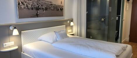 Una habitación de hotel moderna con una cama grande, lámparas de noche, cabina de ducha de vidrio y una foto de un avión colgada en la pared.