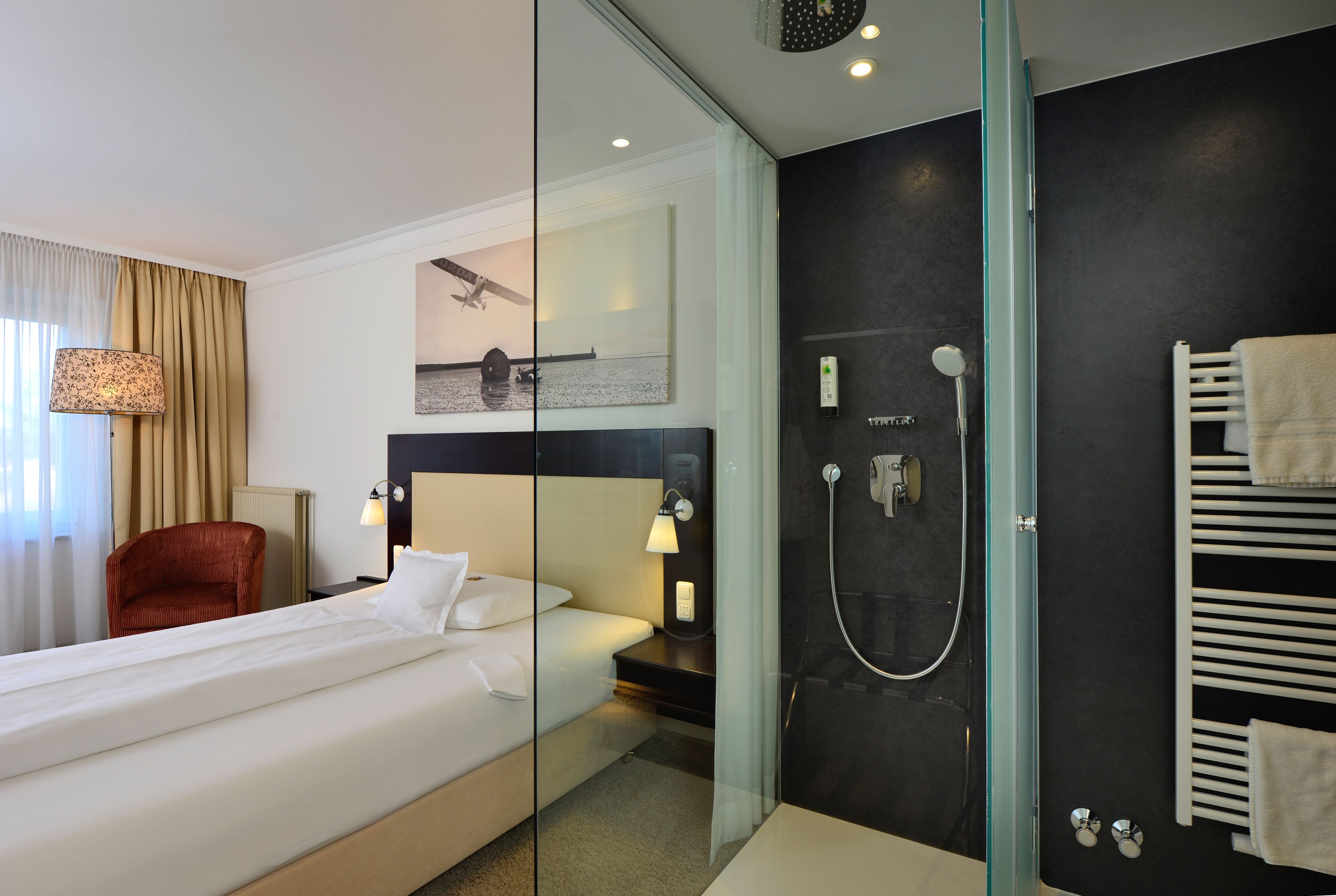 Una habitación de hotel moderna con una cama grande, ducha con cerramiento de vidrio, una silla roja y una obra de arte colgada en la pared.