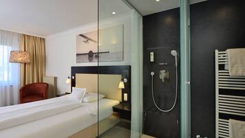 Una habitación de hotel moderna con una cama grande, ducha con cerramiento de vidrio, una silla roja y una obra de arte colgada en la pared.