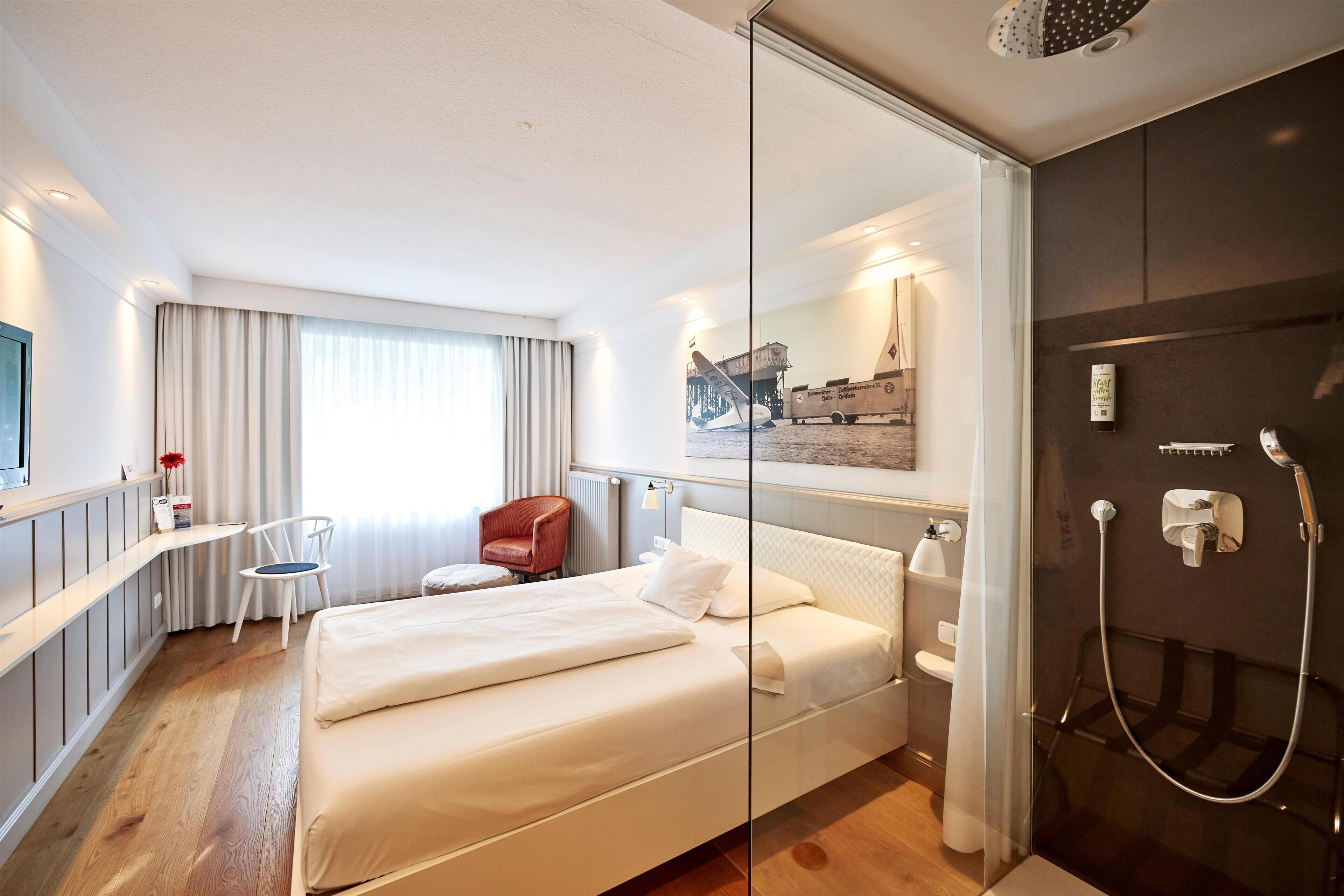 A modern hotel room with a bed, a desk, a chair, a shower, and a large window with curtains.
