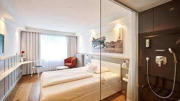 A modern hotel room with a bed, a desk, a chair, a shower, and a large window with curtains.
