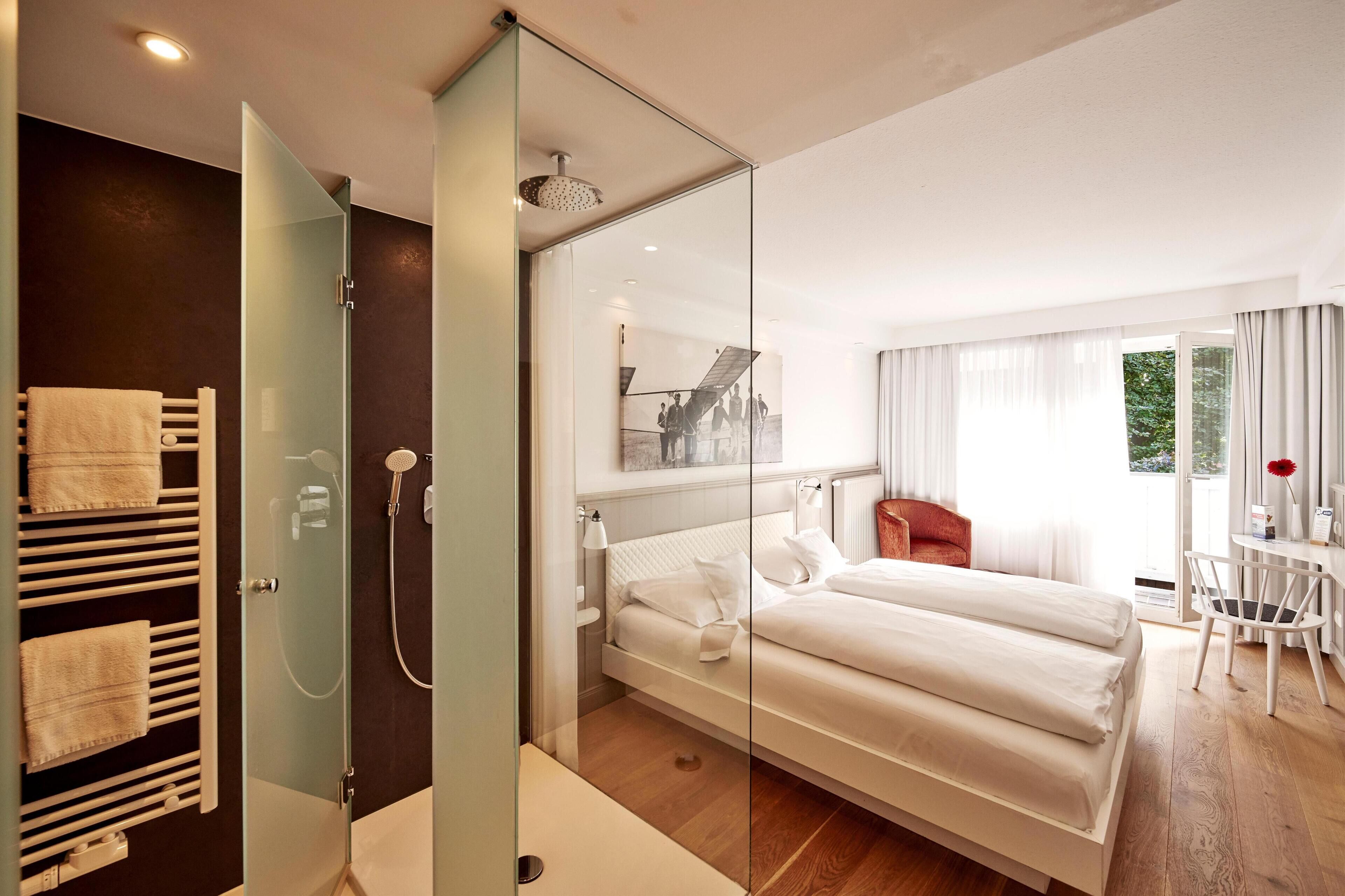 A modern hotel room with a large bed, a glass shower enclosure, a bedside table, and a view of the outdoors.