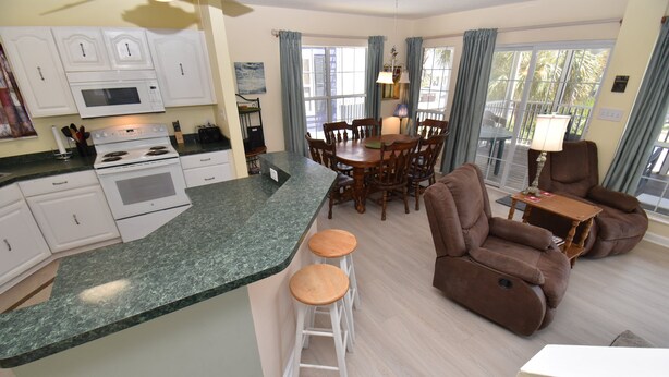 A modern kitchen with white cabinets, a granite countertop, and a dining area with a wooden table and chairs. - Pebble Beach - Second Row in Surfside Beach (Surfside Beach)