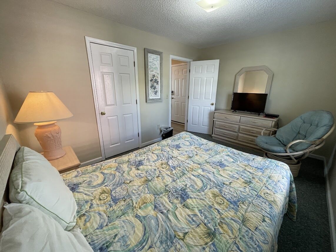 A bedroom with a bed, a chair, a dresser, a lamp, and two doors.