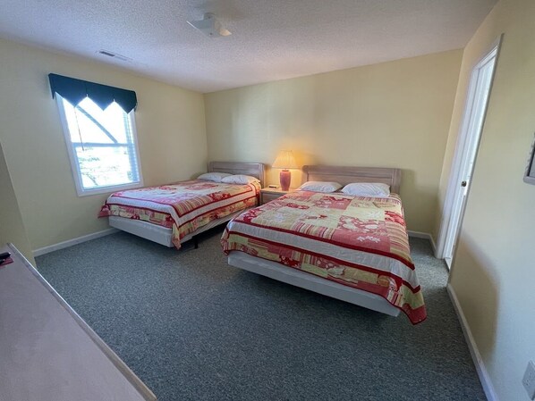 A bedroom with two beds, a nightstand with a lamp, a window with curtains, and a door.