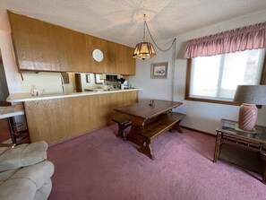 Ett kompakt kök med en träbänkskiva, ett matbord med bänkar, en hängande lampa och ett fönster med persienner.